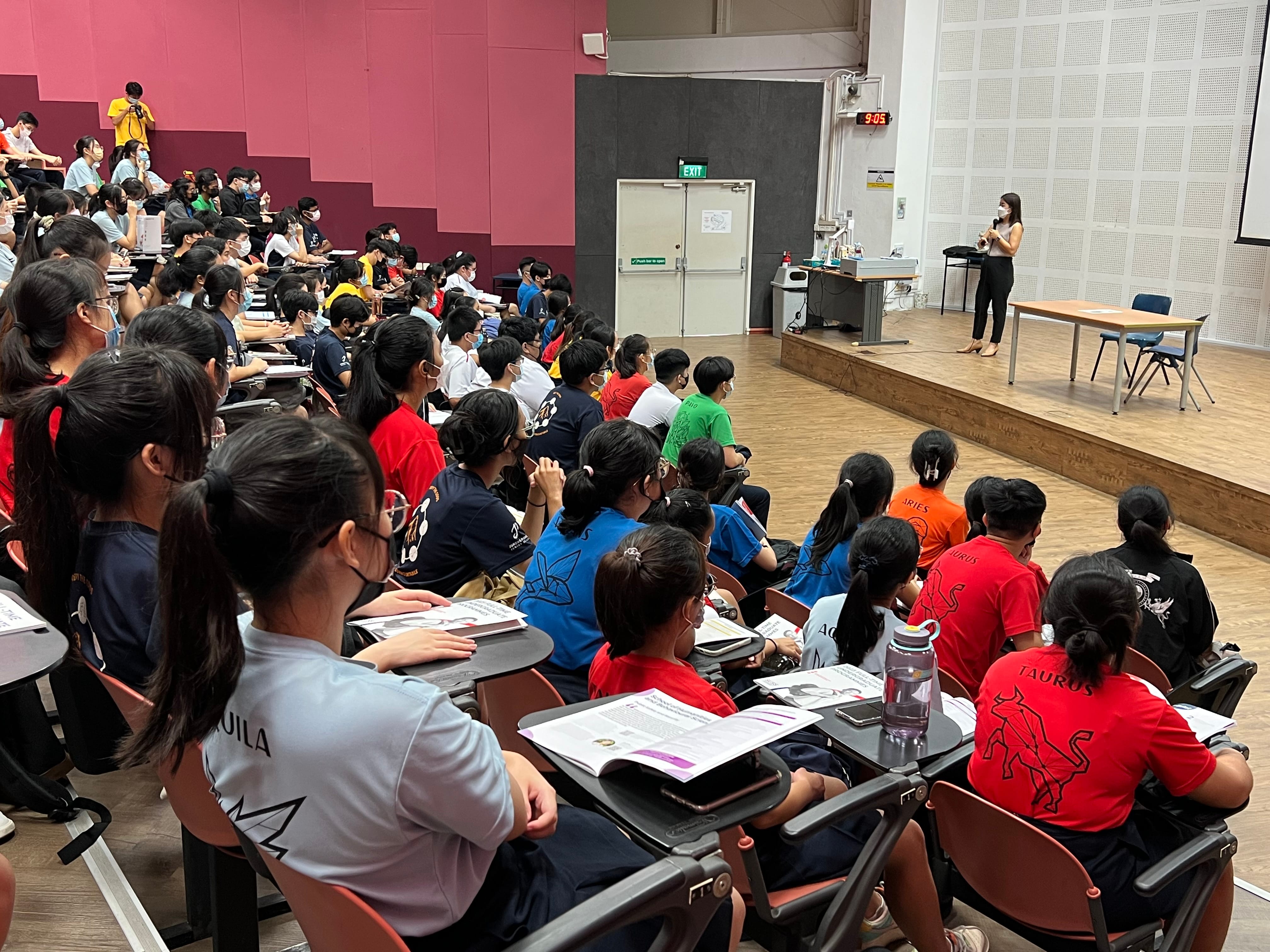 A large lecture hall filled with students at desks facing a female presenter standing on a stage.